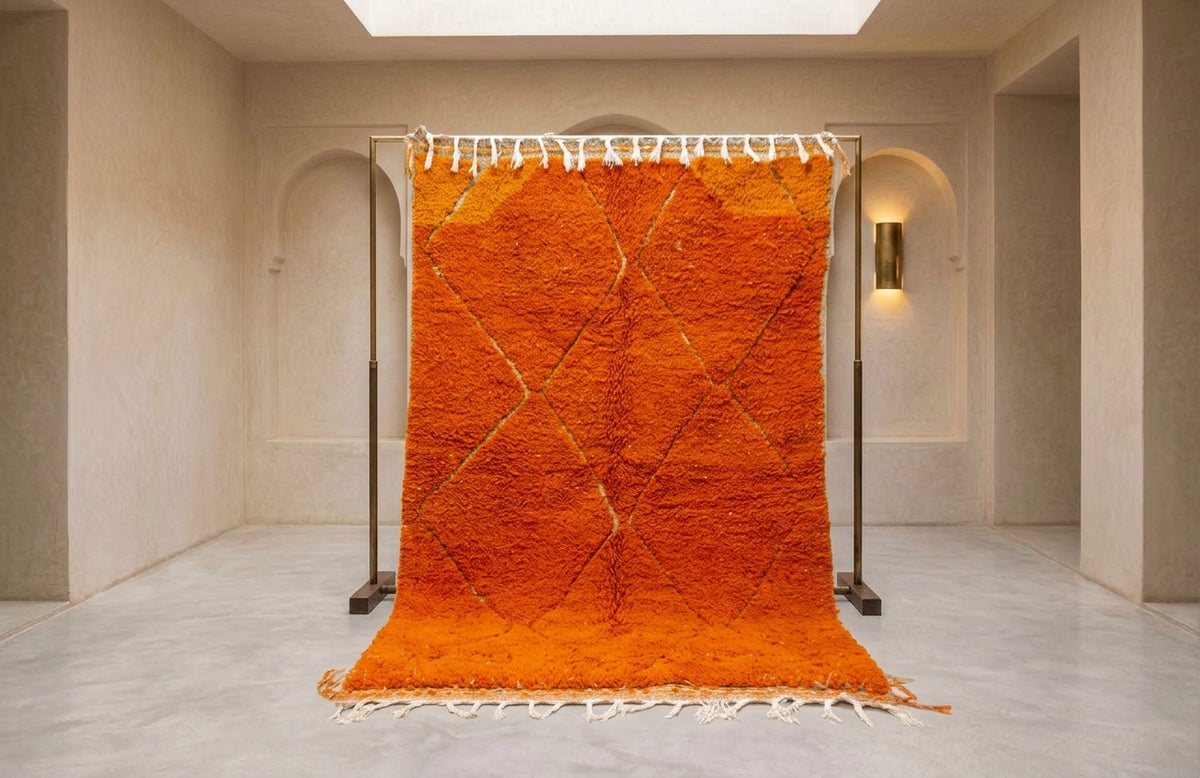 A bright orange Moroccan-style shag rug with white fringe, hanging from a black metal stand in a minimalist beige room.