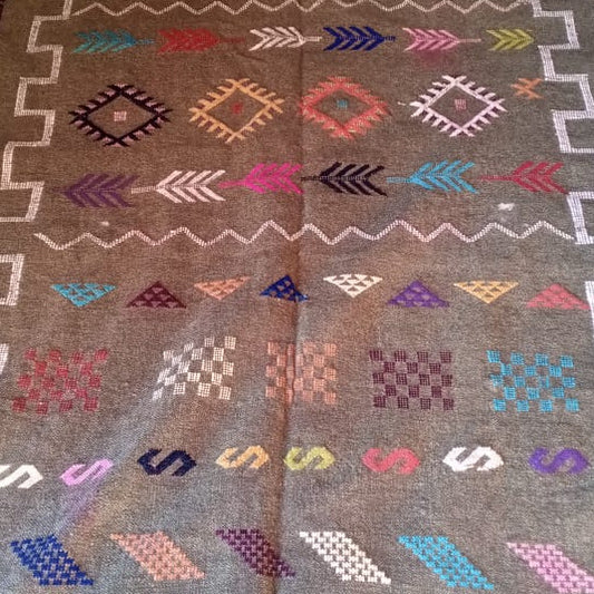 A rectangular woven rug laid flat, featuring a muted gray-green base with white stepped border lines and rows of multicolored geometric and arrow-like motifs. The rug is photographed indoors on a floor with other rugs partially visible around the edges.