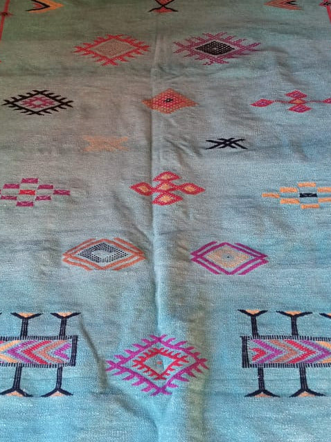 A long turquoise blue wool kilim rug with geometric diamond and tribal patterns in pink, red, black, and beige, laid out on a patterned floor.