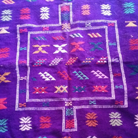 A purple Moroccan wool rug with intricate multicolor geometric patterns and borders, laid flat on a colorful textile surface.