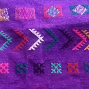 A purple Moroccan-style woven runner rug with colorful geometric patterns, laid flat on top of a larger multicolored patterned rug.