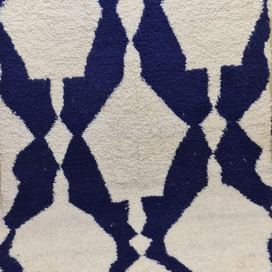 A rectangular woven rug with a bold geometric diamond pattern in dark navy blue and cream, featuring fringed edges, displayed on a tiled floor against a beige wall.