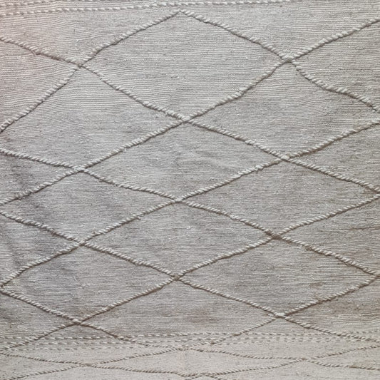 A natural beige woven rug with a diamond lattice pattern and fringed edges, displayed on a tiled floor with other rugs in the background.