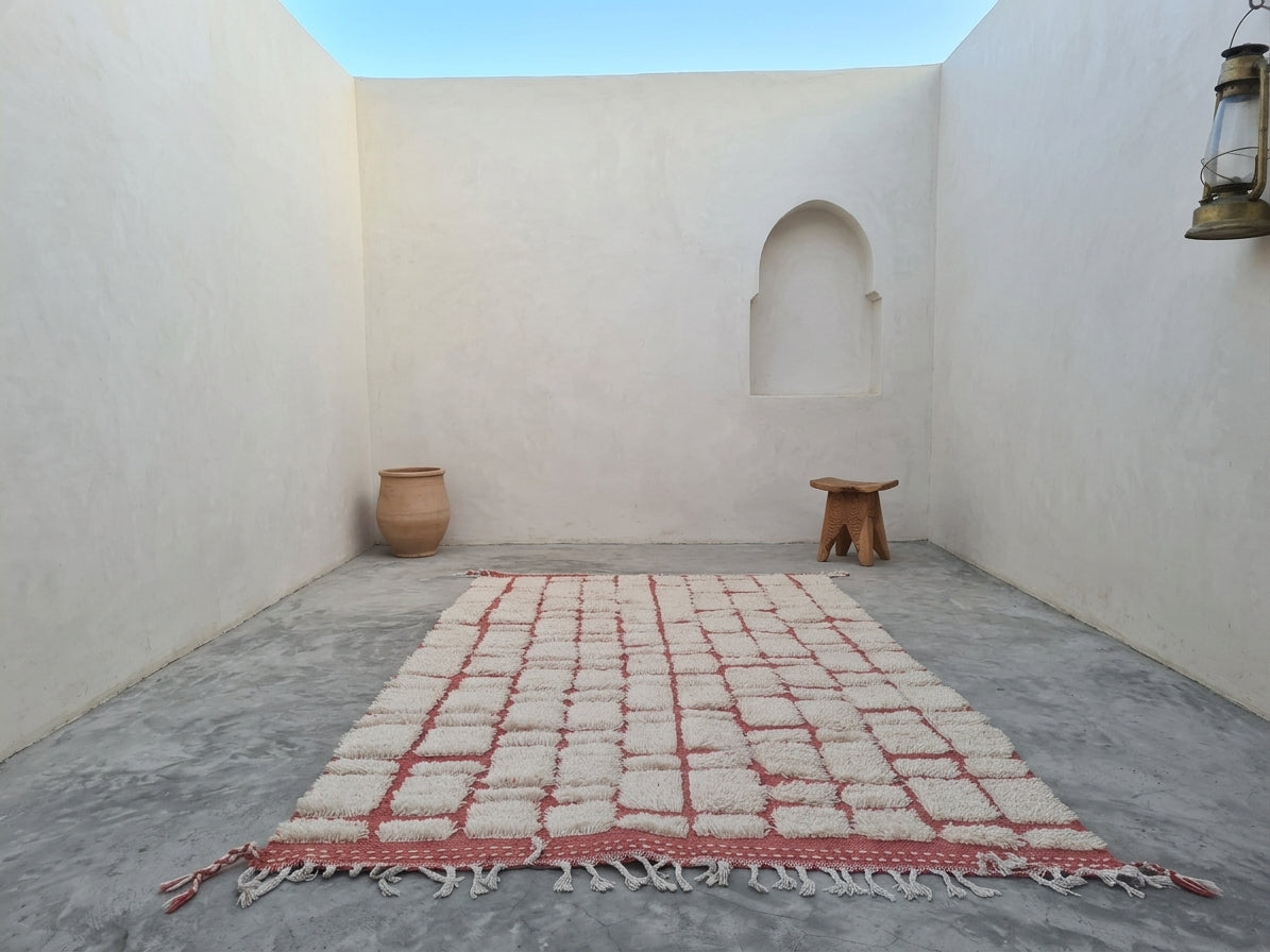 A rectangular Moroccan-style woven rug with a cream and terracotta red pattern and fringe tassels, displayed on a gray concrete floor in a minimalist white-walled courtyard.