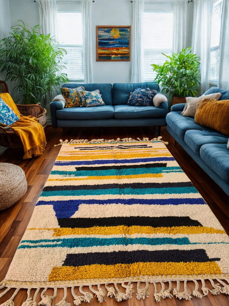 Boujaad rug in mustard, navy, and ivory horizontal bands, styled in a living room with blue velvet sofas and leafy indoor plants.