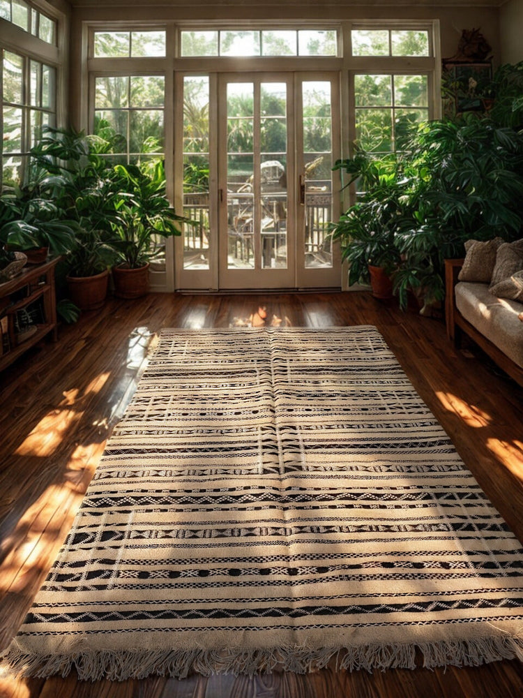 Striped Moroccan kilim rug in neutral tones, placed in a sunlit room with large windows and indoor greenery.