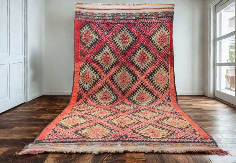 Ait Zayan rug in saturated red, pink, and saffron diamond patterns, displayed upright against a white wall and dark wood floor.