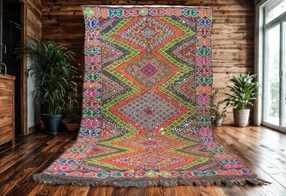 Colorful patterned rug on a wooden floor with plants and a wooden cabinet in the background.