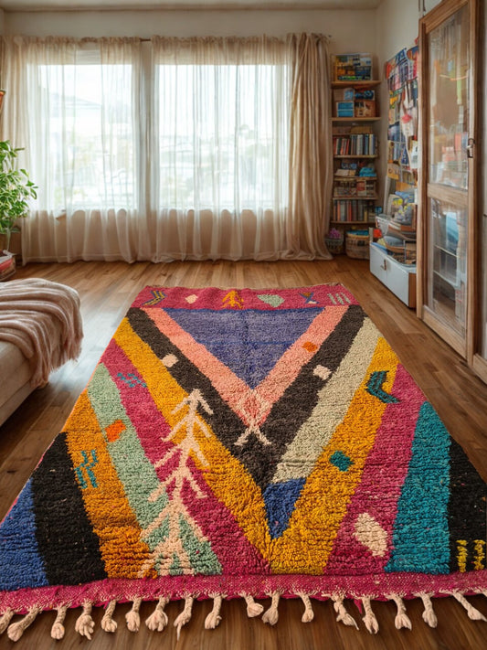 Colorful geometric-patterned rug on a wooden floor with a bookshelf and window in the background.