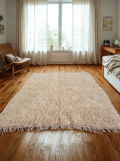 Beige shaggy rug on a wooden floor with a chair and window in the background