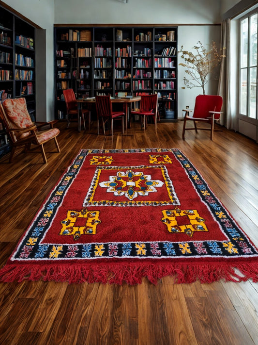 Decorative rug with colorful patterns on a wooden floor in a room with bookshelves and chairs.