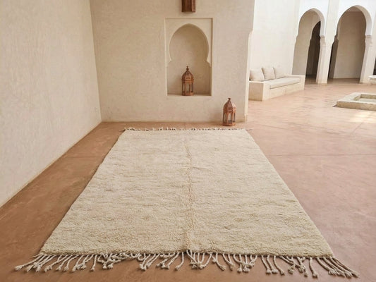 A large cream-colored shag rug with fringe tassels laid out on a terracotta tiled floor in a Moroccan-style room with arched alcoves and built-in seating.