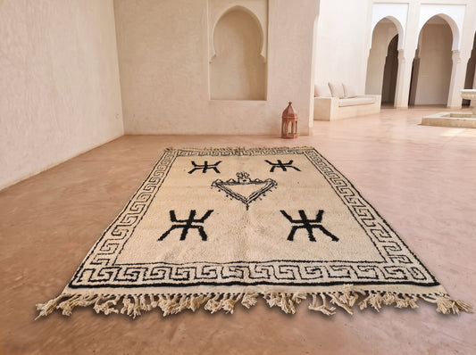A rectangular woven rug with a cream base and black geometric motifs, including a Greek key border and central crown emblem, displayed on a terracotta-colored floor in a Moroccan-style courtyard with white stucco walls and arches.