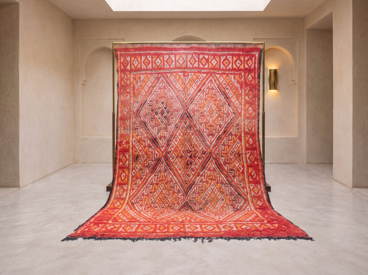 A large patterned area rug displayed hanging from a frame in a minimalist beige gallery space with arched walls and a light floor.