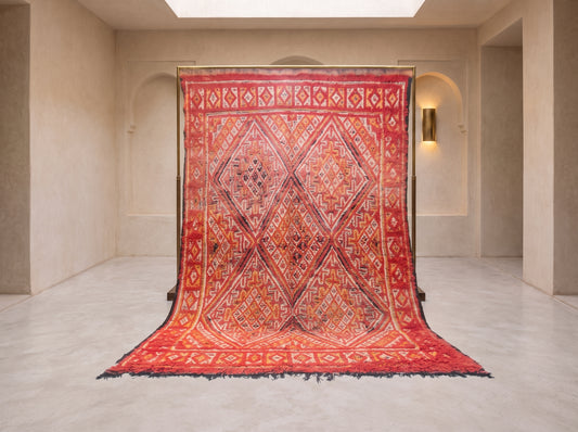 A large patterned area rug displayed hanging from a frame in a minimalist beige gallery space with arched walls and a light floor.