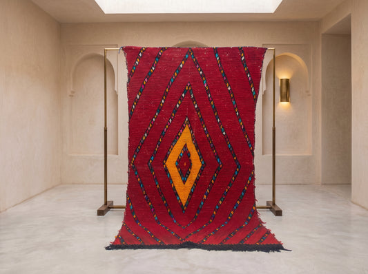 A large red Moroccan-style woven rug with a bold geometric diamond pattern in yellow, orange, blue, and black, displayed on a minimalist brass stand in a warm, cream-colored gallery space.