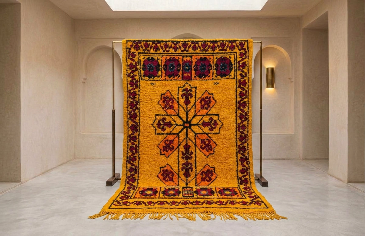 A large yellow wool rug with a central cross motif and red, black, and blue geometric borders, hanging from a metal stand in a minimalist beige gallery space.