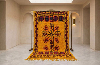 A large yellow wool rug with a central cross motif and red, black, and blue geometric borders, hanging from a metal stand in a minimalist beige gallery space.