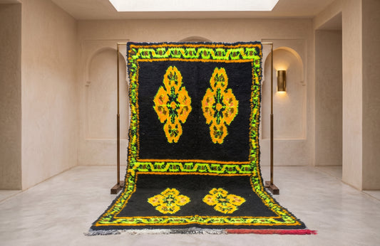 A large black wool rug with vibrant yellow and green geometric patterns, displayed on a wooden stand in a minimalist beige gallery space.