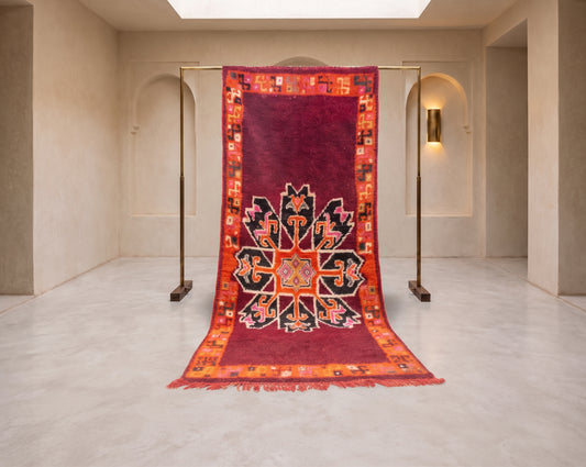 A large traditional oriental rug with a deep red center and a colorful geometric border, hanging from a metal display stand in a minimalist beige gallery space.
