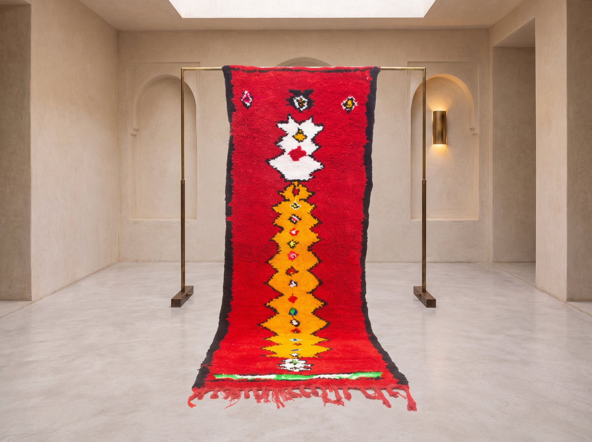 A vibrant red Moroccan-style wool rug with a central yellow and white geometric pattern, hanging from a minimalist brass stand in a modern, minimalist gallery space with beige walls and arched doorways.