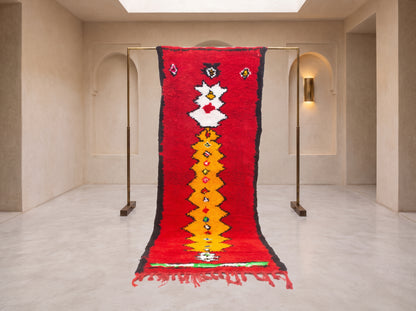 A vibrant red Moroccan-style wool rug with a central yellow and white geometric pattern, hanging from a minimalist brass stand in a modern, minimalist gallery space with beige walls and arched doorways.