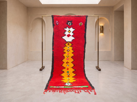 A vibrant red Moroccan-style wool rug with a central yellow and white geometric pattern, hanging from a minimalist brass stand in a modern, minimalist gallery space with beige walls and arched doorways.