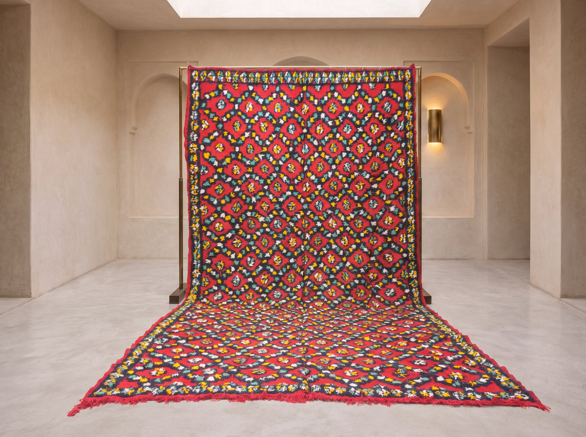 A large patterned rug or textile panel displayed upright in a minimalist beige gallery space, with its lower portion draped forward. The textile features a dense multicolor floral and geometric motif dominated by red, with black, yellow, blue, green, and white throughout.