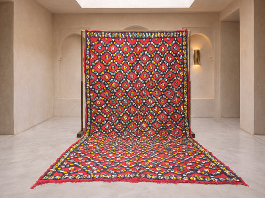 A large patterned rug or textile panel displayed upright in a minimalist beige gallery space, with its lower portion draped forward. The textile features a dense multicolor floral and geometric motif dominated by red, with black, yellow, blue, green, and white throughout.