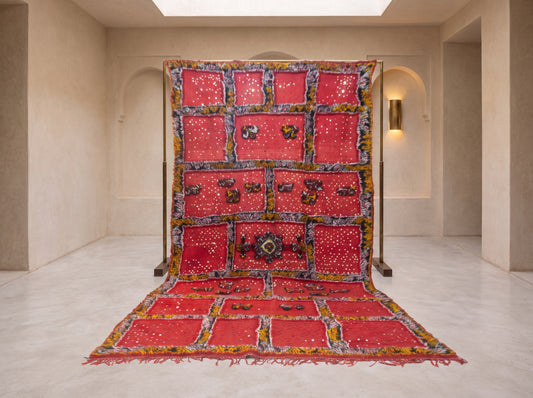 A large red textile artwork or rug panel displayed on a stand in a minimalist beige gallery space, with matching red panels draped below. The textile features multiple rectangular sections, decorative borders, and small reflective embellishments.