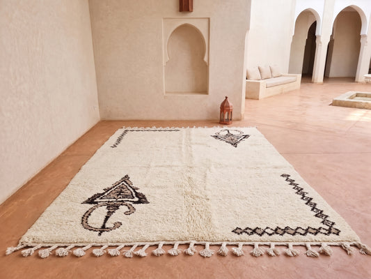 A large off-white Moroccan-style rug with black geometric and tribal patterns, displayed on a terracotta tiled floor in a Moroccan courtyard with beige walls and arches.