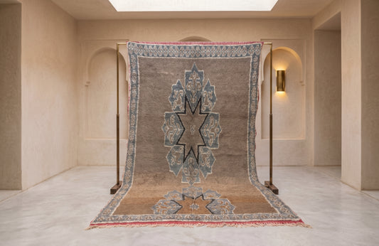A large traditional oriental rug displayed hanging from a wooden stand in a minimalist beige room with arched walls and a skylight.