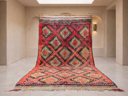 A large kilim-style area rug with a bold geometric diamond pattern, displayed hanging from a rod in a minimalist beige room.