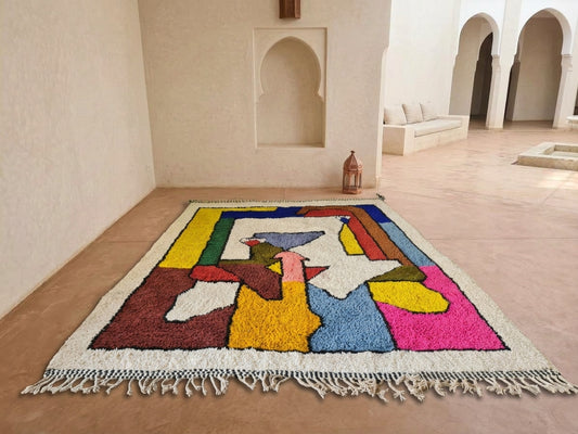 A colorful Moroccan-style area rug with a geometric abstract pattern and white fringe, displayed on a terracotta tiled floor in a minimalist Moroccan courtyard.