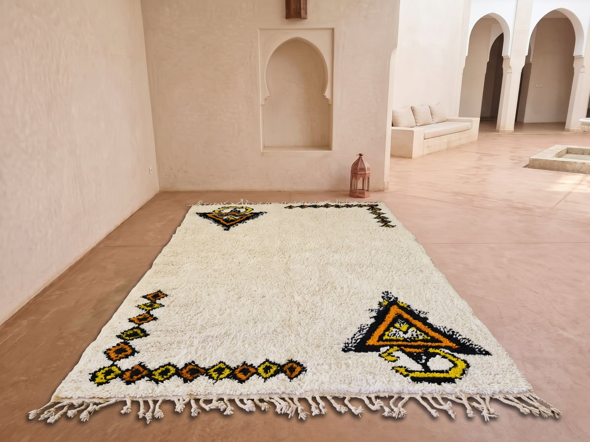 A large off-white Moroccan wool rug with colorful geometric patterns and tassels, displayed on a terracotta tiled floor in a minimalist, sunlit courtyard with arched alcoves.