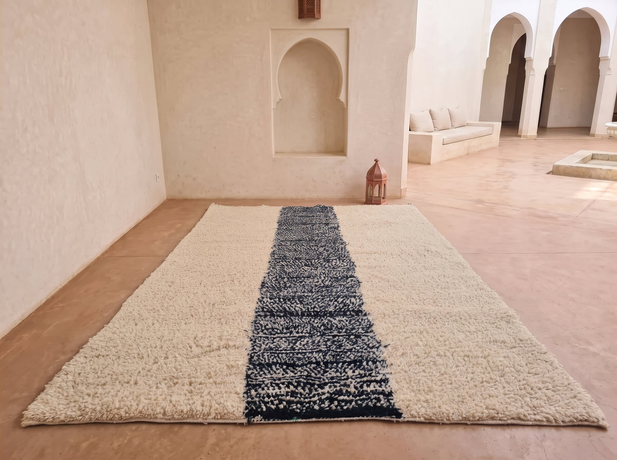 A large rectangular area rug laid out in a sunlit Moroccan courtyard. The rug has a thick, shaggy cream-colored border with a central stripe of dark navy blue and cream woven pattern.
