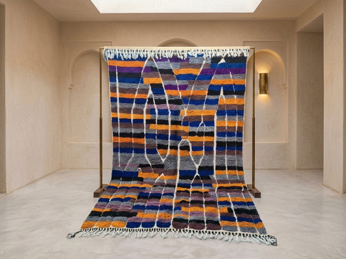 A large Moroccan-style wool rug displayed on a wooden stand in a minimalist beige room. The rug features horizontal stripes in navy blue, orange, purple, black, and white, with vertical white fringe-like patterns running down the center.
