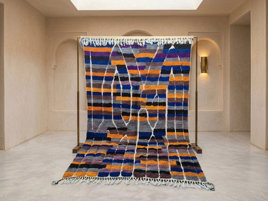 A large Moroccan-style wool rug displayed on a wooden stand in a minimalist beige room. The rug features horizontal stripes in navy blue, orange, purple, black, and white, with vertical white fringe-like patterns running down the center.