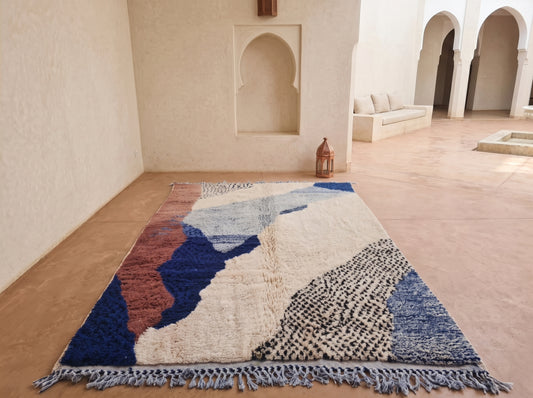 A rectangular area rug with an abstract, hand-woven Moroccan design in shades of blue, terracotta, cream, and black, displayed on a terracotta floor in a minimalist courtyard setting.