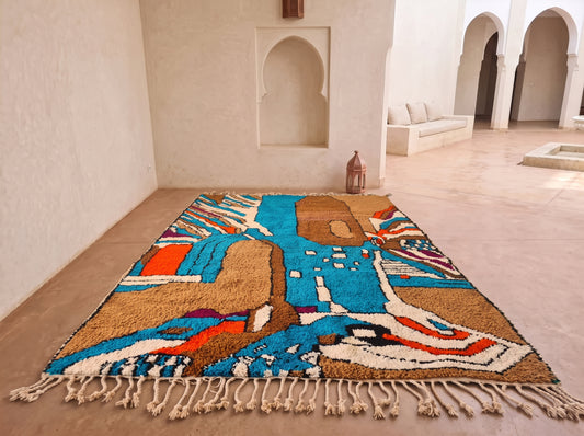 A rectangular handwoven rug with a colorful abstract geometric pattern and cream fringe, displayed on a tiled floor in a Moroccan-style courtyard with arched alcoves.