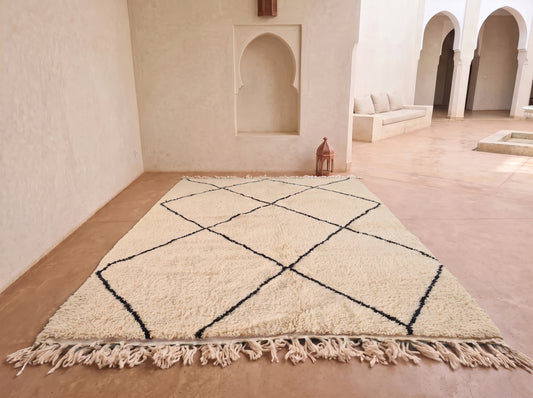 A large cream-colored Moroccan-style rug with black geometric diamond lines and fringe, displayed on a terracotta floor in a minimalist, sunlit room with white walls and arches.