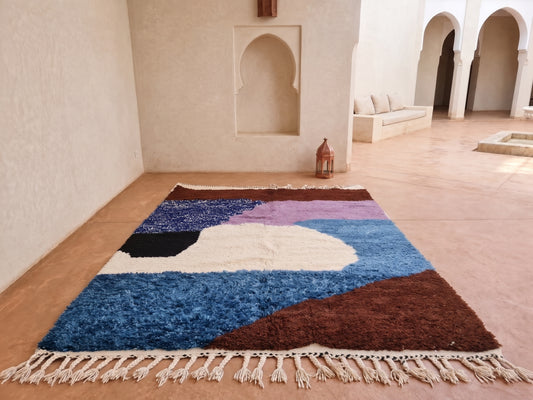 A rectangular shag area rug with cream fringe, featuring an abstract geometric pattern in shades of blue, brown, purple, black, and cream, displayed on a terracotta floor in a Moroccan-style courtyard.