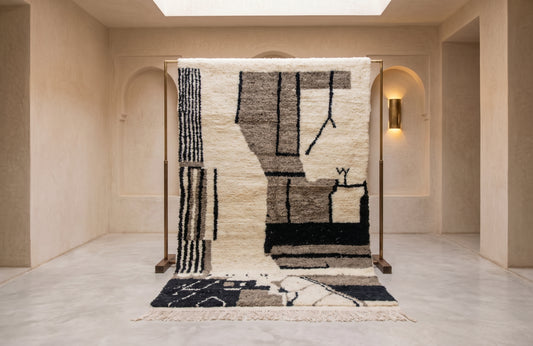 A large abstract area rug displayed on a wooden stand in a minimalist beige gallery space. The rug features a geometric pattern in black, charcoal, and cream.