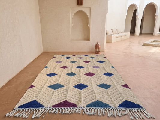 A rectangular Moroccan-style area rug with a cream base and geometric diamond patterns in blue, purple, and teal, laid out on a terracotta floor in a minimalist Moroccan courtyard.