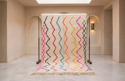 A large woven area rug displayed on a metal stand in a minimalist beige interior space. The rug has a cream base with vertical zigzag stripes in black, pink, red, blue, orange, and yellow, finished with a fringed edge.