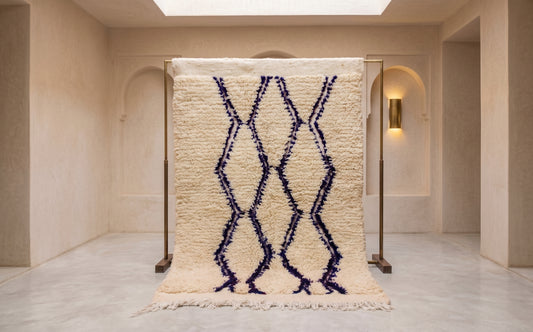 A high-pile textured Moroccan-style rug in cream with a dark navy blue geometric diamond pattern, displayed on a wooden stand in a minimalist beige interior room.