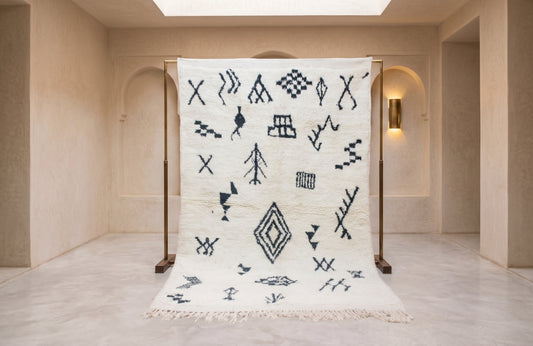 A large white Moroccan-style rug with black geometric patterns is displayed on a wooden stand in a minimalist beige interior room.
