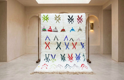 A large white woven rug with colorful geometric and abstract patterns, displayed on a wooden stand in a minimalist beige gallery space.