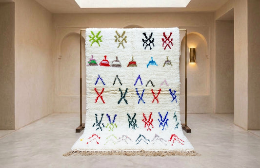A large white woven rug with colorful geometric and abstract patterns, displayed on a wooden stand in a minimalist beige gallery space.