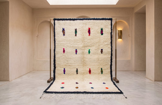 A large cream-colored Moroccan-style shag rug displayed on a wooden stand in a minimalist beige gallery space. The rug features a thick black border, small colorful geometric motifs, and a fringed bottom edge.